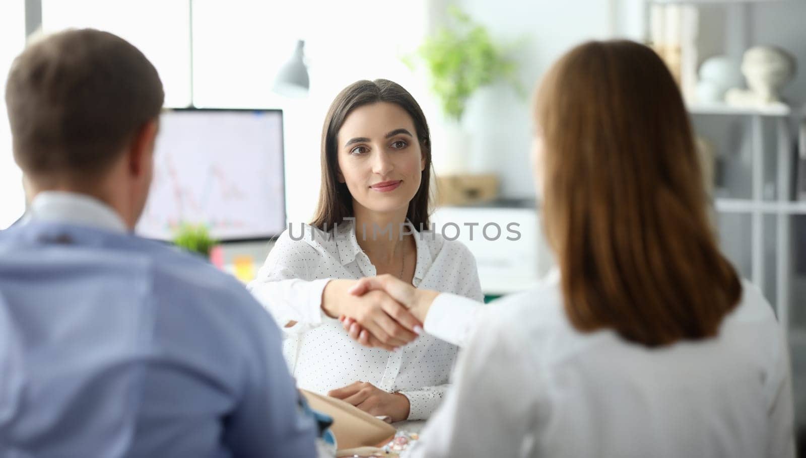 Portrait of joyful businesswoman sitting at modern workplace and shaking hand with woman partner. Businesslady looking at partner with calmness. Business negotiations concept