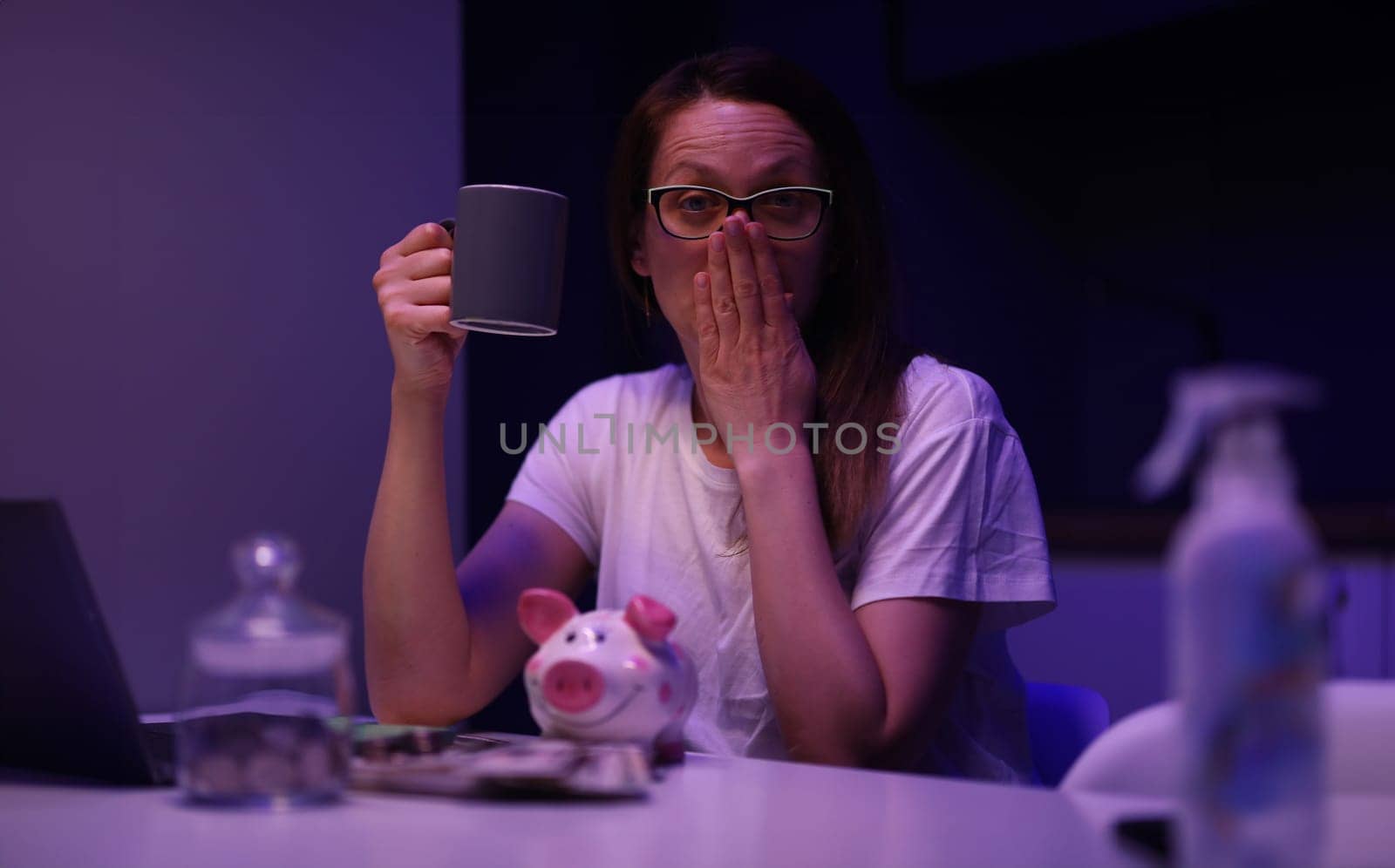 Portrait of tired young woman yawning sitting in room with cup of coffee and accounting money from piggy bank. Saving up for future. Finance and family budget concept