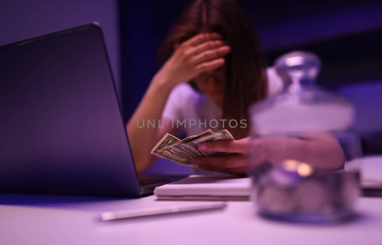 Close-up of depressed woman calculating family financial situation. Person close face with hand and holding banknotes. Glass container with coins. Money and investment concept