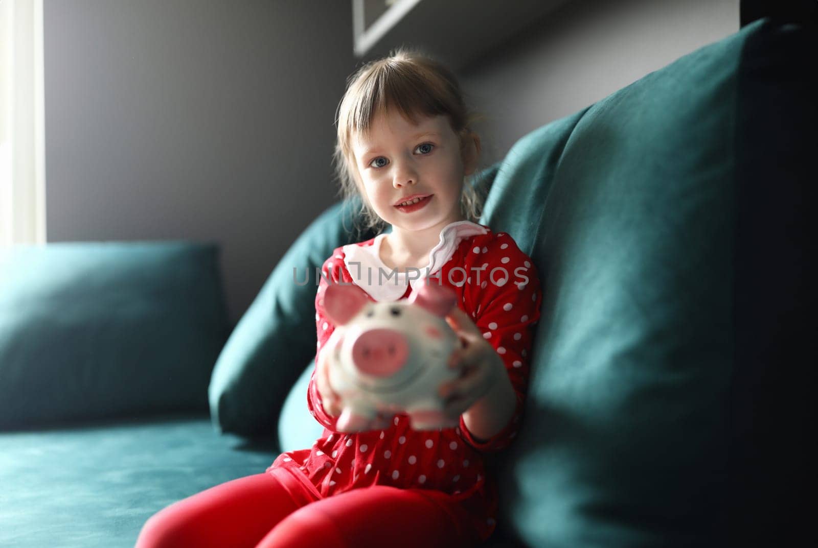 Portrait of smiling beautiful child holding piggy bank sitting on sofa indoors. Lovely blonde kid saving up for future. Girl in red dress. Childhood and happiness concept