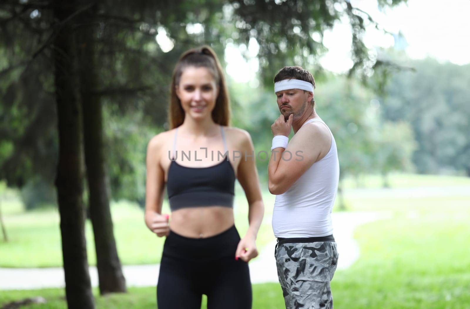 Portrait of middle-aged man checking young beautiful woman on morning run in park. Attractive female in stylish sportswear. Outdoors workout in forest. Sport and hobby concept