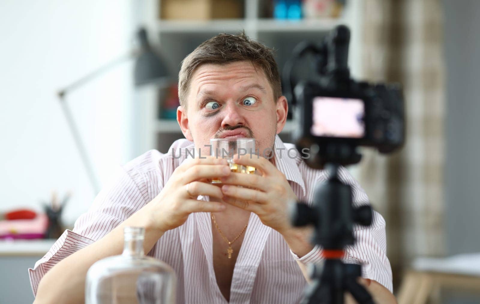 Portrait of drunk male filming on camera smelling glass of whiskey. Middle-aged man making funny face and look messy. Tasting of alcoholic drink on videocamera. Blogger concept