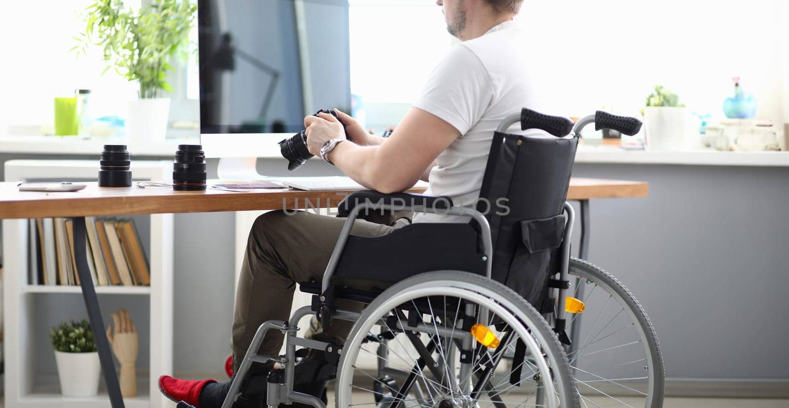 Close-up of professional man photographer holding camera and sitting in wheelchair. Modern office with laptop on desktop. Disabled people and photography business concept