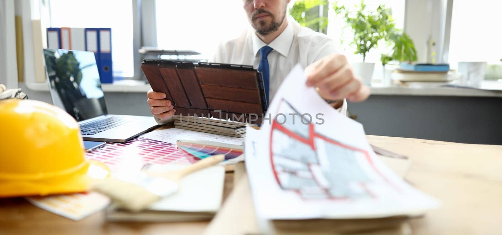 Close-up of creative person in presentable suit working in renovation agency. House plan and palette with colourful sample on wooden table. Interior design and professional architect concept