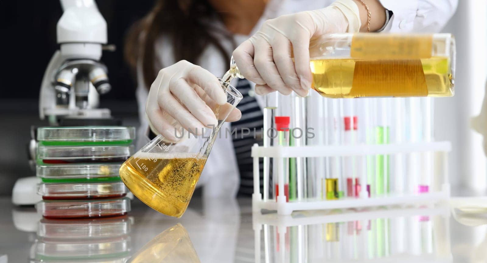 Close-up of laboratory worker pouring alcohol liquid in glass flask. Woman working in protective gloves and uniform. Chemistry and research. Modern investigation and science concept