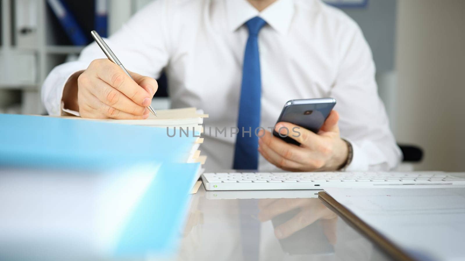 Close-up of businessman filling important document sitting on modern office. Man in presentable suit holding smartphone. Signing contract. Business and technology concept