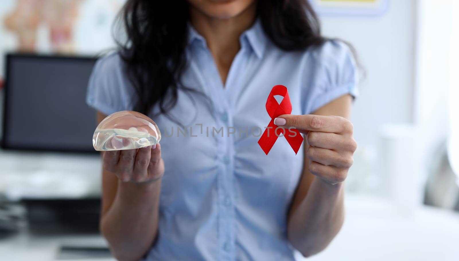 Close-up of female holding red ribbon cancer symbol on one hand. Silicone breast implant on other hand. Confidence and beauty. Modern medicine and womans body concept