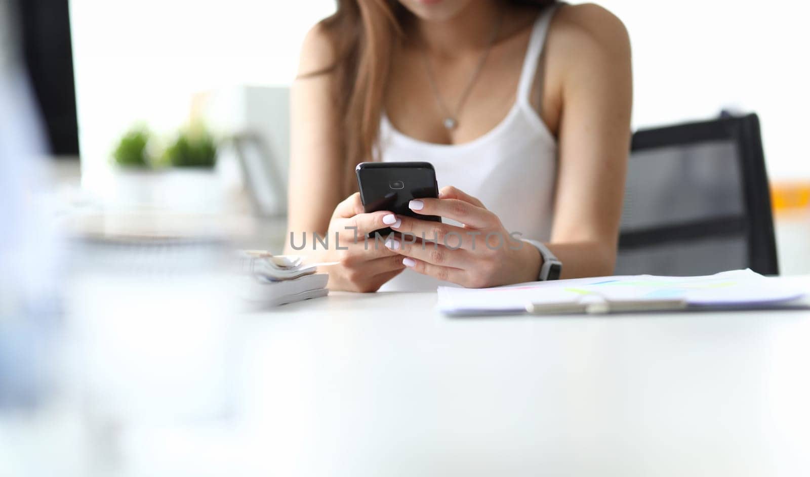 Focus on woman arms holding modern high-tech telephone. Smart lady sitting indoors and typing something on screen of mobile phone. Office concept. Blurred background