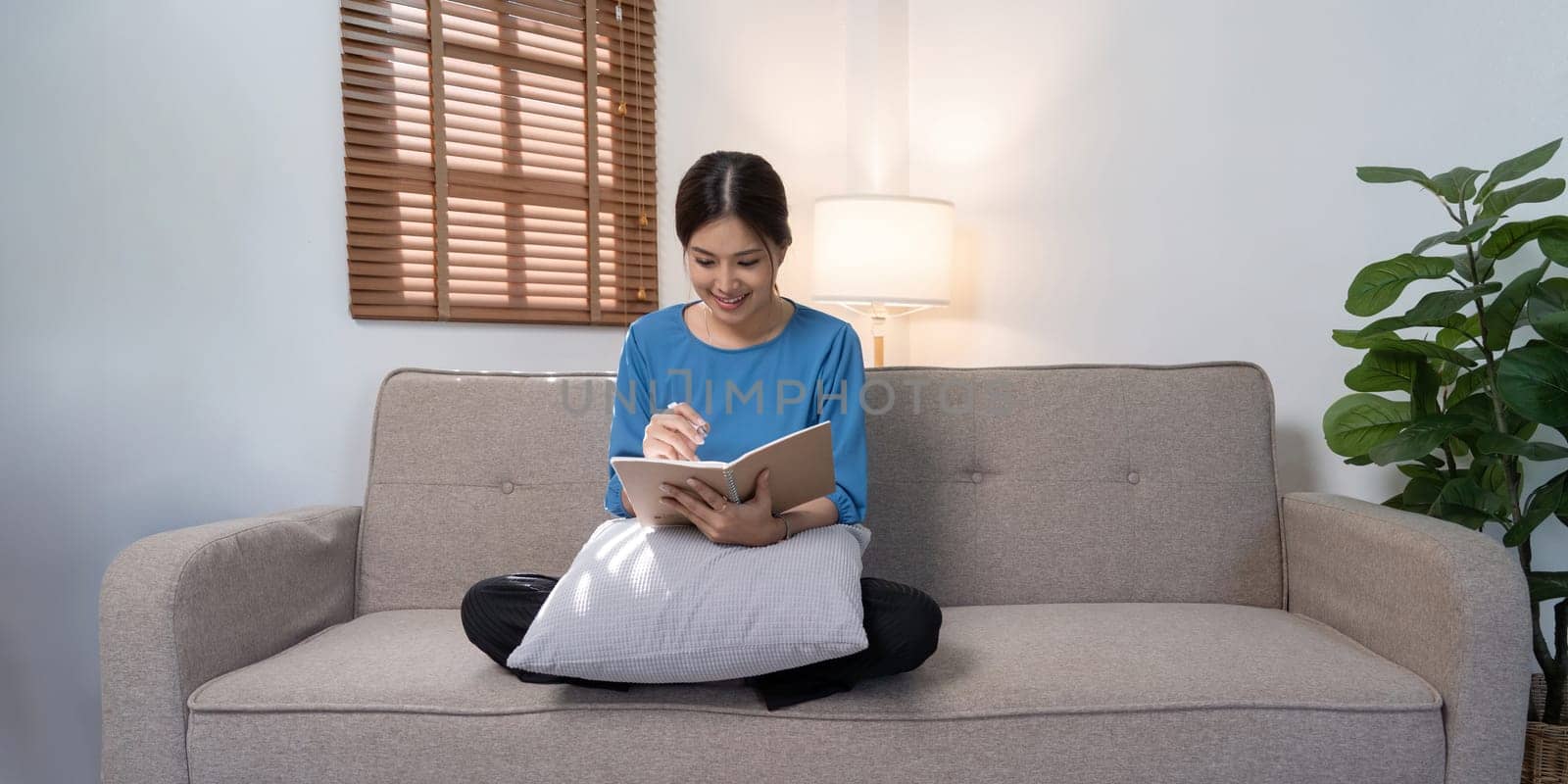 Beautiful young asian woman sitting and taking note on sofa at house. Happy smiling working online, Freelance work at home.