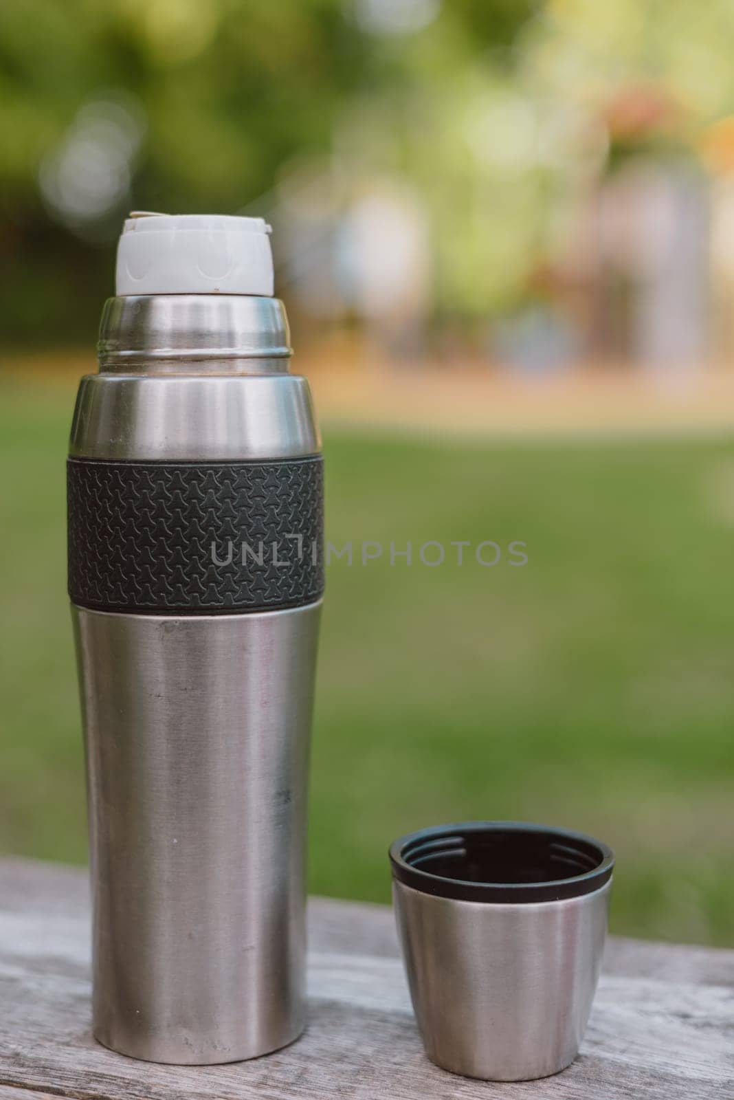 Stainless steel cup, mug with thermos stands on a wooden table in the forest on a green natural background. Thermos and mug with hot drink standing on wet wooden table after rain outdoors, close-up. Steam rises from thermo mug. by Andrii_Ko