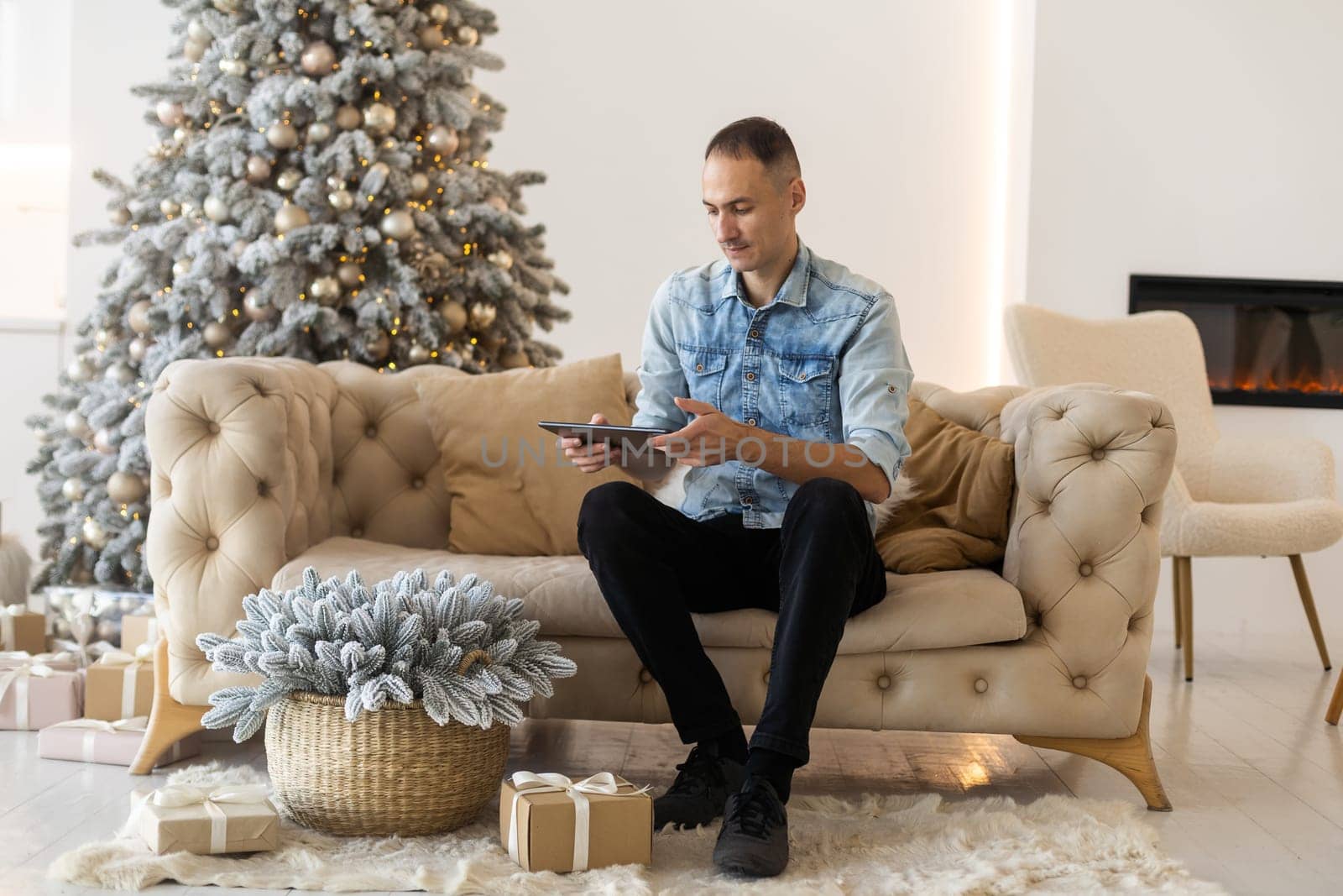 A portrait of young man with tablet sitting on sofa at home at Christmas time. He has video call with family. by Andelov13