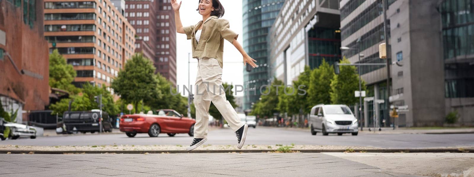 Portrait of asian happy girl jumping and dancing in city centre, posing on streets, express joy and excitement. Copy space