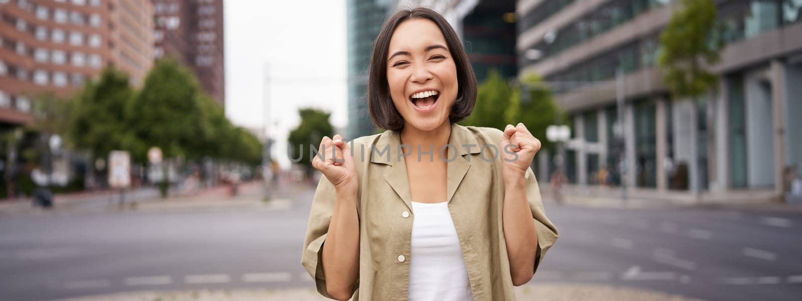 Happy asian girl triumphing on streets of city, dancing from happiness, celebrating victory, posing outdoors by Benzoix