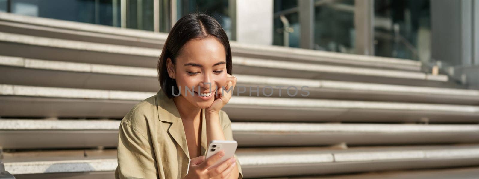 Vertical shot of asian woman, student sits on stairs in city, looking at mobile phone screen and smiling, using smartphone app.