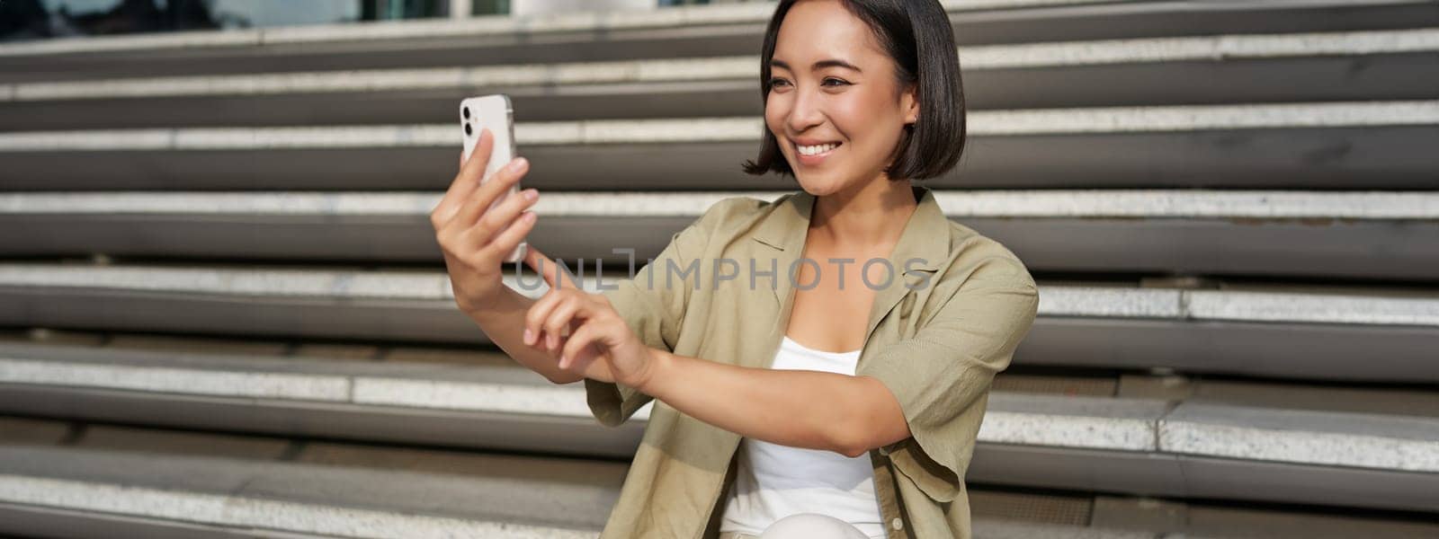 Portrait of asian beautiful girl takes selfie on smartphone. Young korean woman sits on stairs outdoors and makes photos on mobile phone.