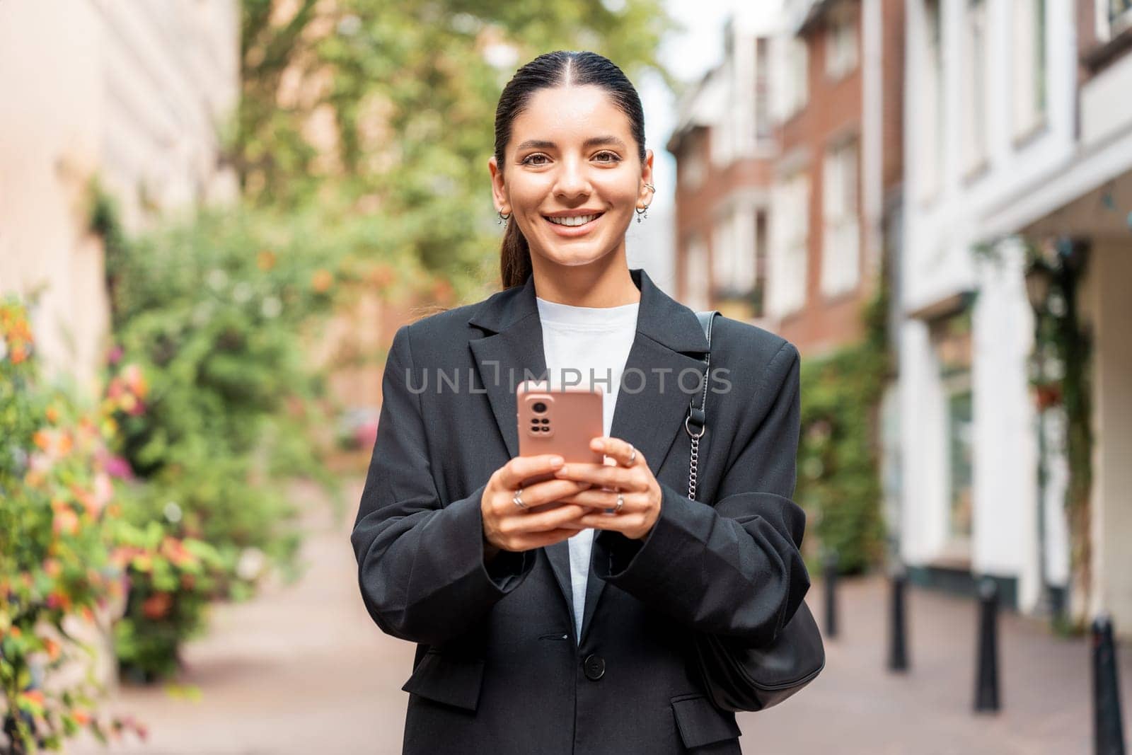 Sunny portrait of a newly entrepreneur young woman in her 25s, black suit, holding a phone smiling, looking in camera by AndreiDavid