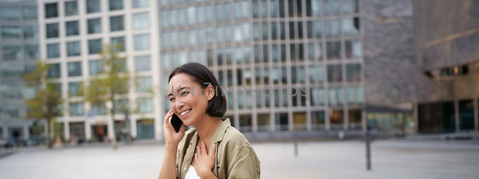 Vertical shot of smiling stylish asian girl, having a telephone conversation and walking on street. Young woman talking on mobile phone.