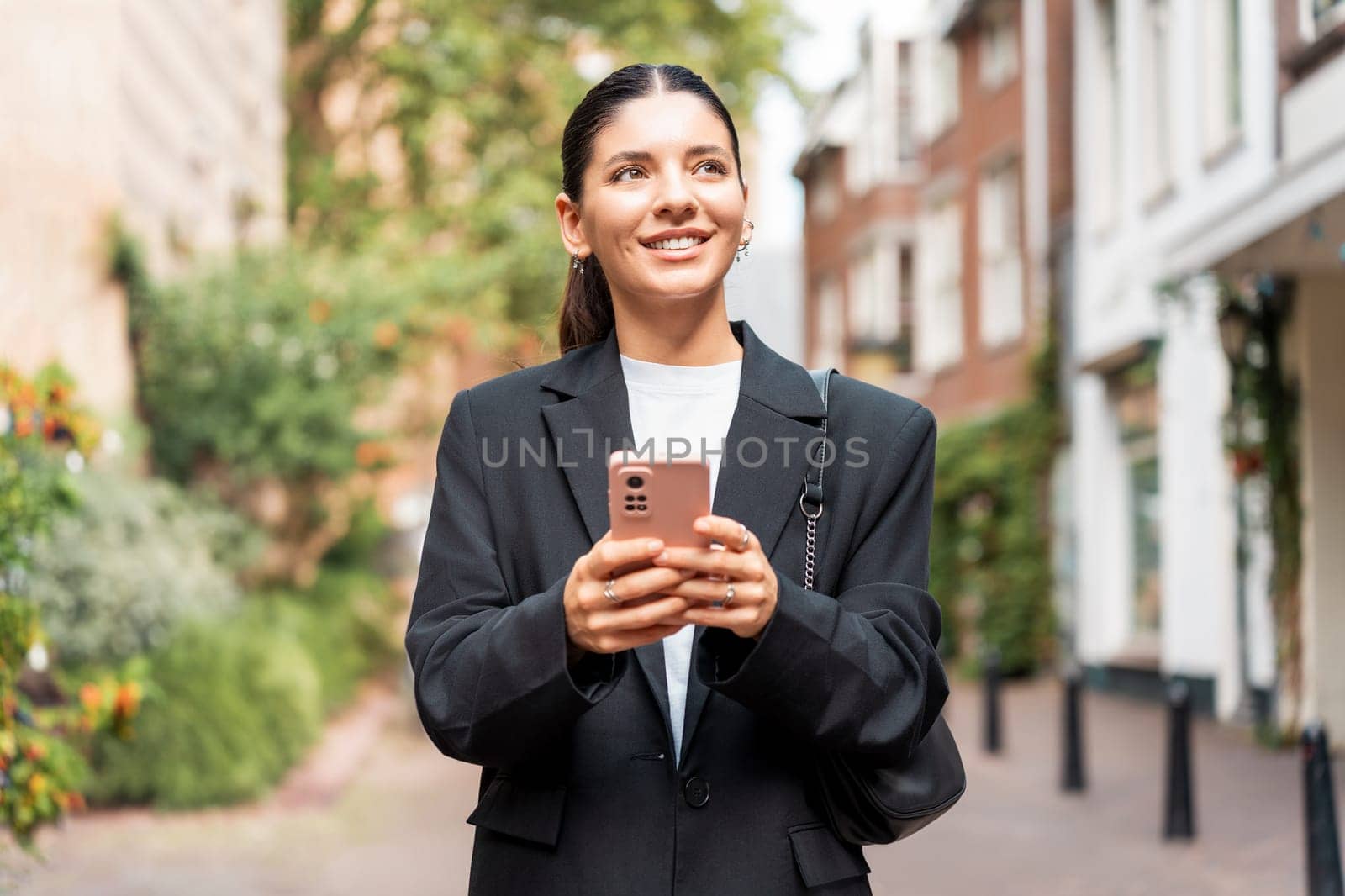 Sunny portrait of a newly entrepreneur young woman in her 25s, black suit, holding a phone smiling, looking away.