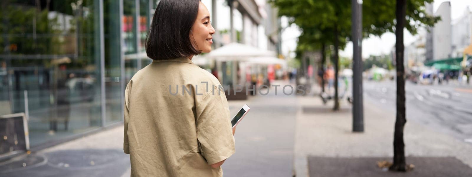 Rear shot of young woman walking in city, going down the street and smiling, holding smartphone. View from behind.