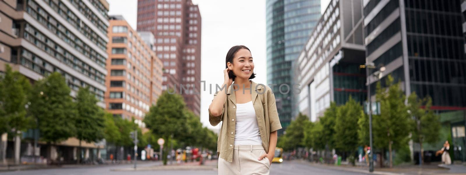 Outdoor shot of young smiling asian woman, standing on street in daytime, looking around, posing happy.