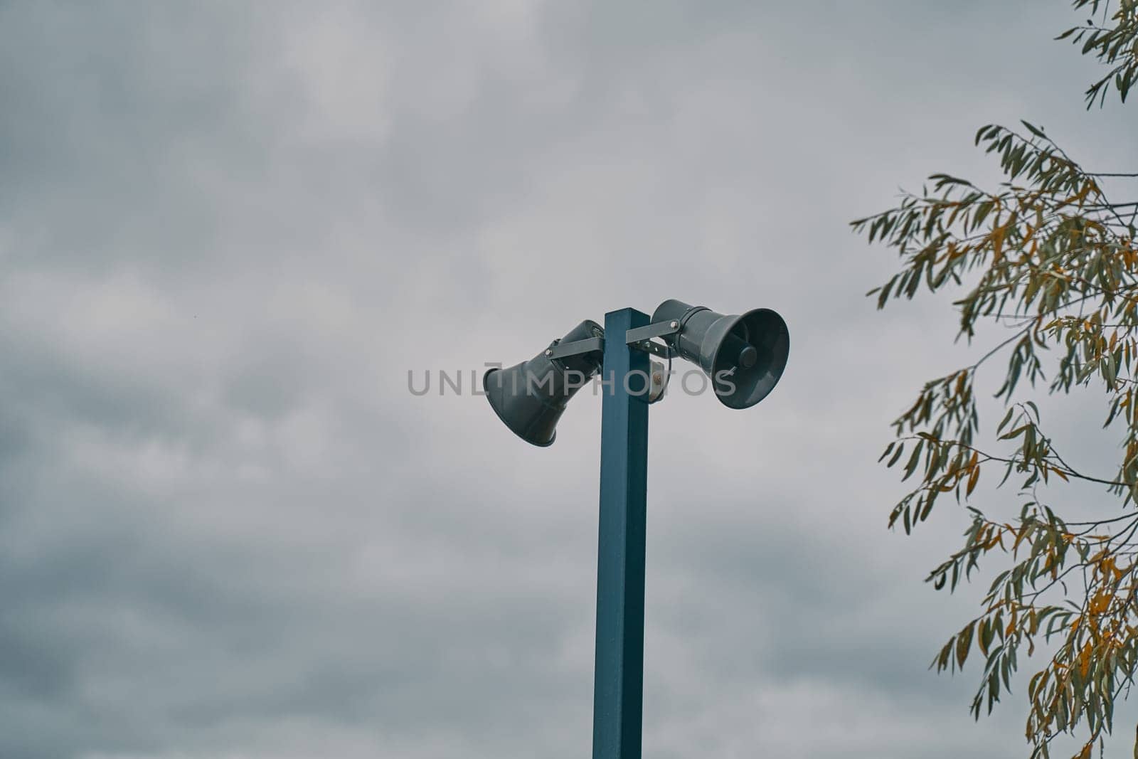 Tall metal column with two gray loudspeakers against cloudy sky. Hazard warning system. Providing security in the city, notification of emergencies. Copy space for text.