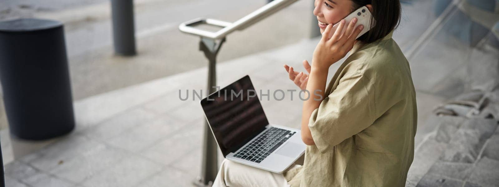 Smiling asian woman makes a phone call. Girl student using laptop and mobile phone, talking to someone on telephone.