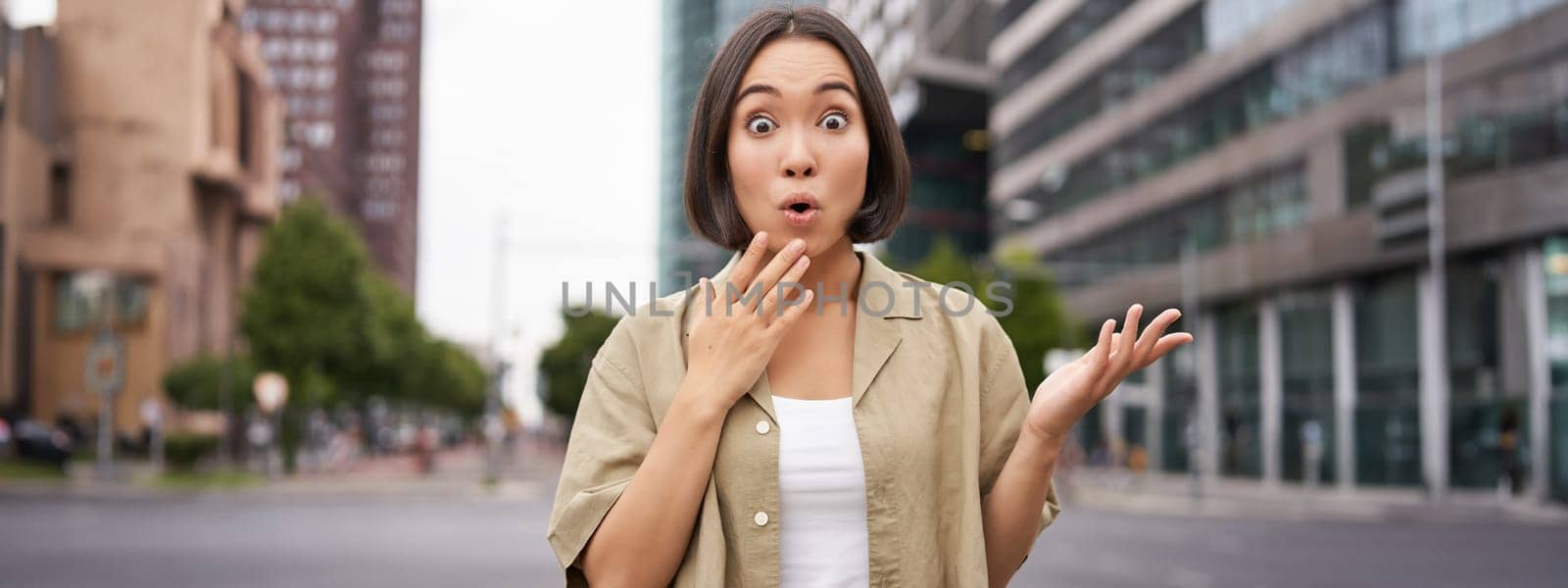 Modern asian girl standing on street with surprised face, looking impressed and amazed, hear awesome news by Benzoix