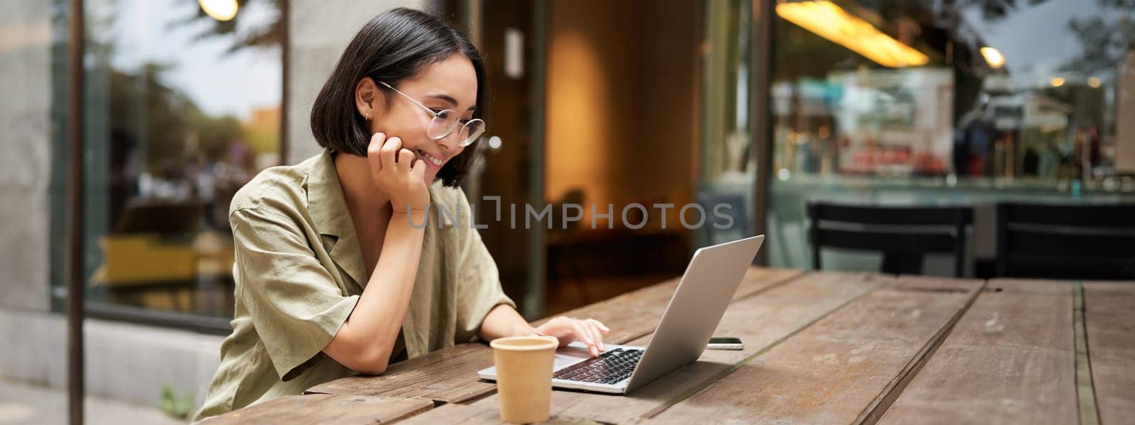 Portrait of smiling girl in glasses, sitting with laptop in outdoor cafe, drinking coffee and working remotely, studying online by Benzoix