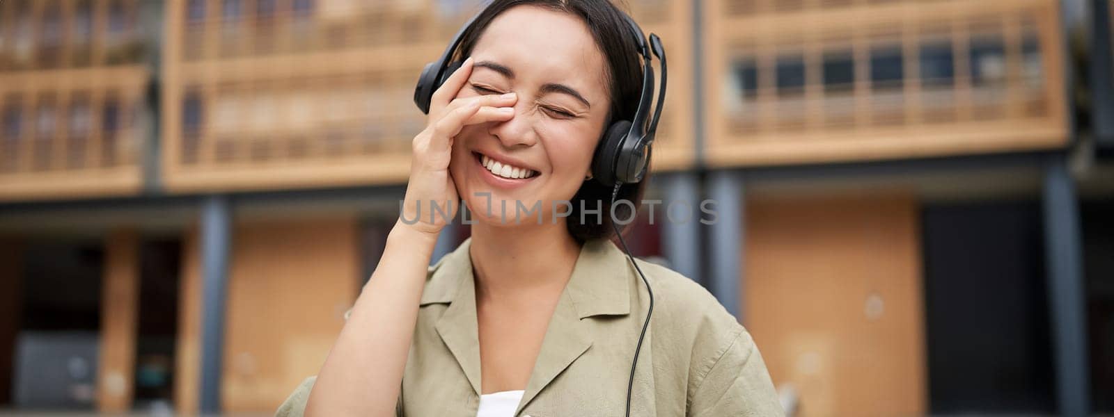 Portrait of beautiful asian woman in headphones, listening music on street of city centre, smiling happily.