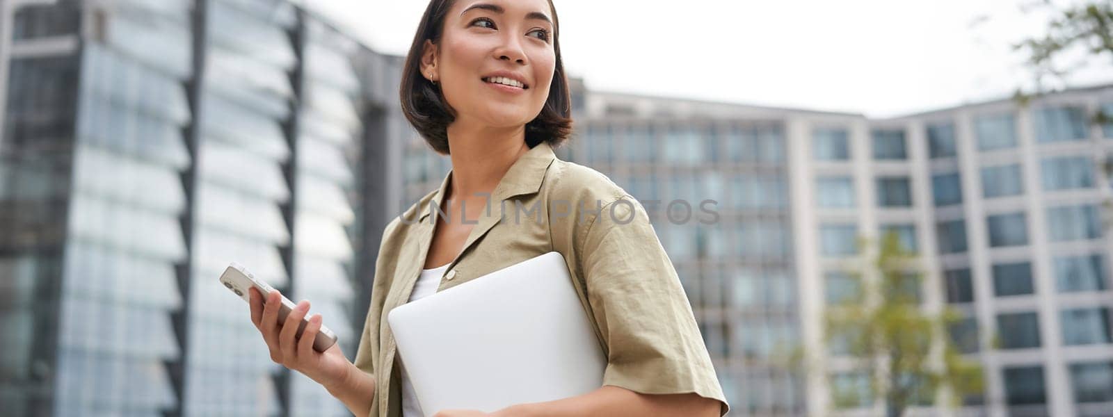 Portrait of asian girl with laptop, going to work or uni, holding smartphone and smiling, walking on street.