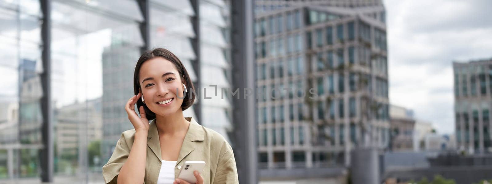 Young smiling asian woman in wireless earphones, walks on street with smartphone and headphones by Benzoix