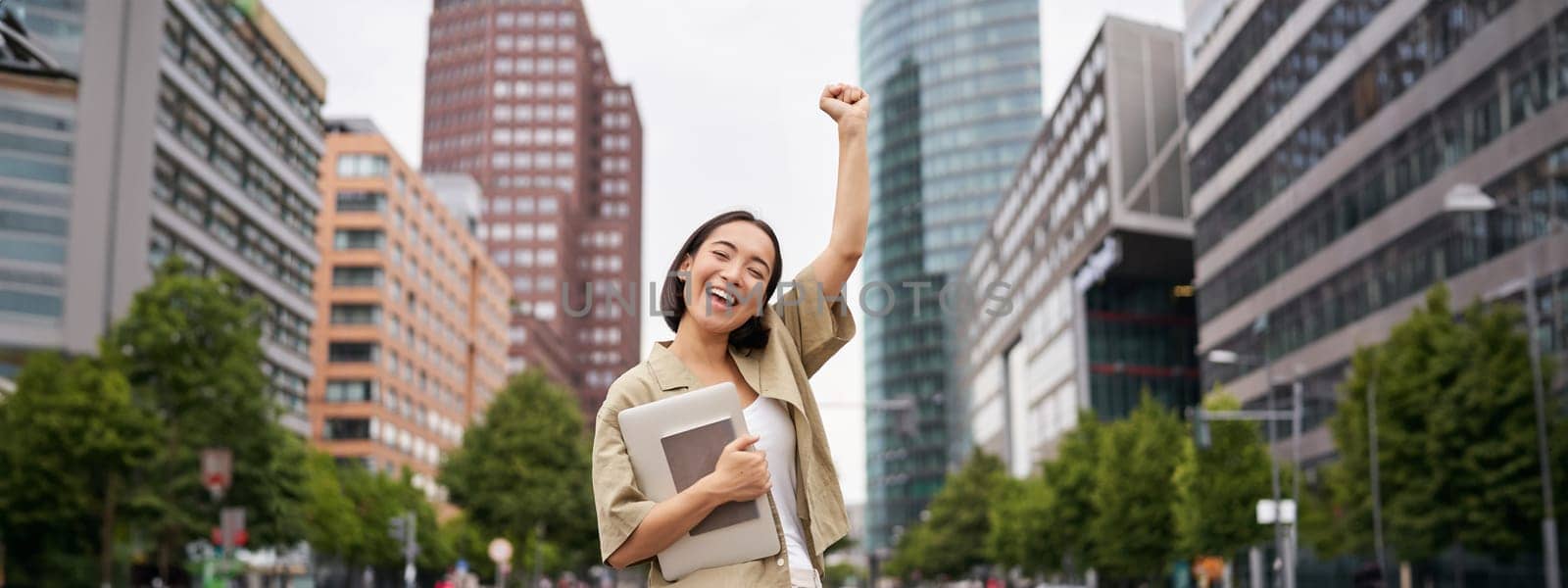 Portrait of happy asian woman stands with tablet near street road, cheering, raising hand up in triumph, celebrating.