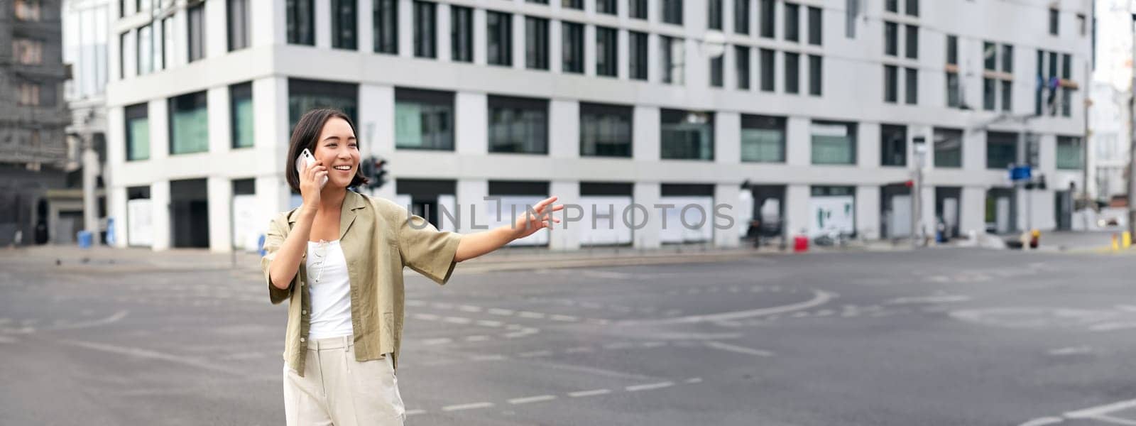 Young woman waving to taxi driver, calling or talking on mobile phone, order car ride, standing outdoors on street.