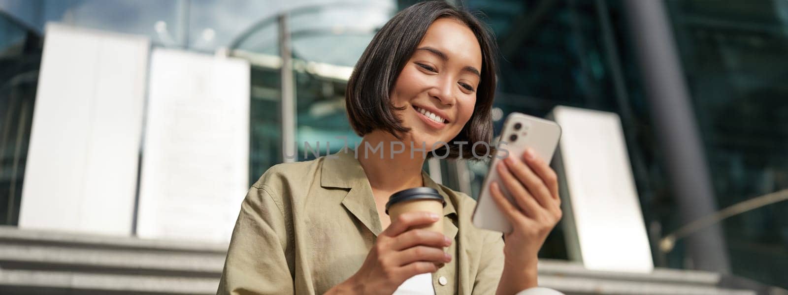 Beautiful smiling asian girl, drinking coffee, using mobile phone and sits on stairs outside. Young woman video chat outdoors.