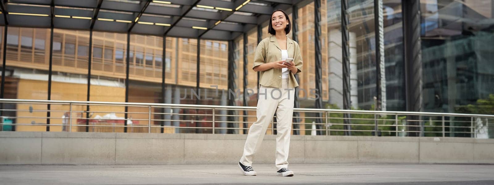 Portrait of young asian woman, student with smartphone, standing on street near glass building, waiting for someone by Benzoix