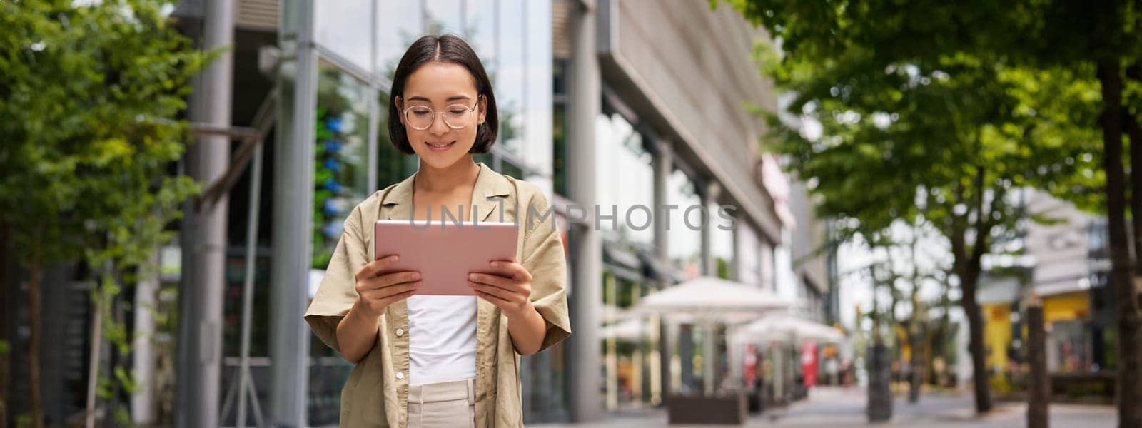 Outdoor shot of young korean woman stands on street with laptop, wears glasses, reads, smiles happily.