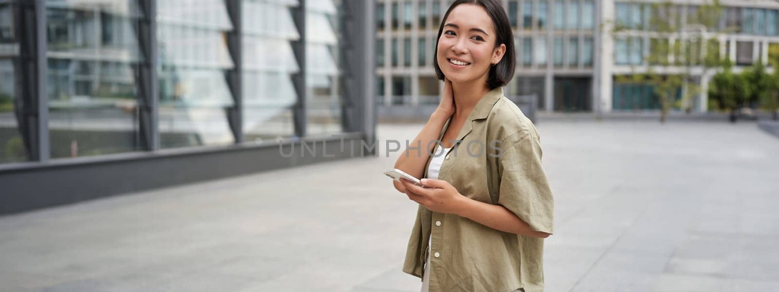 Portrait of asian woman walking in city. Street style shot of girl with smartphone, posing outdoors on street.