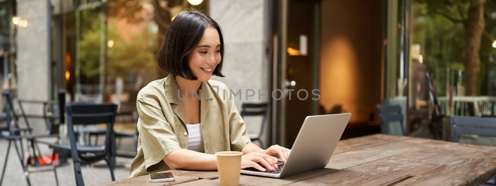 Young asian woman, digital nomad working remotely from a cafe, drinking coffee and using laptop, smiling.