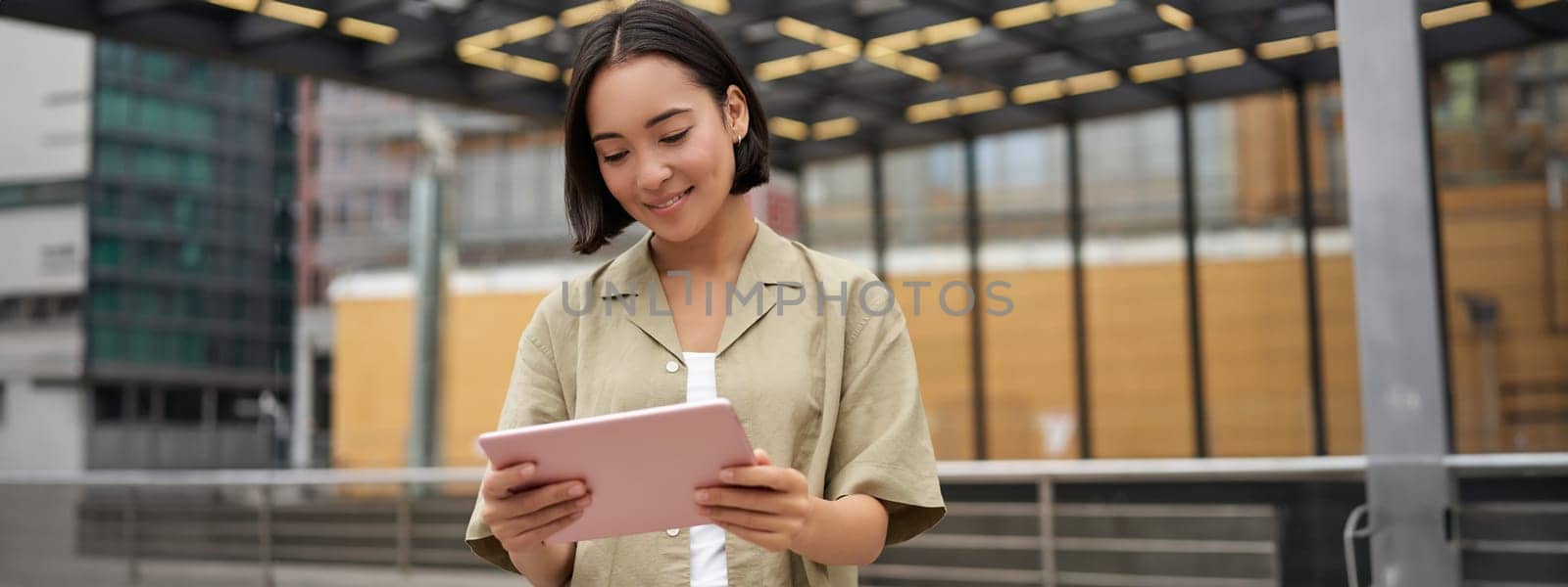 Portrait of beautiful asian girl using tablet, reading, looking at tablet while standing on street.