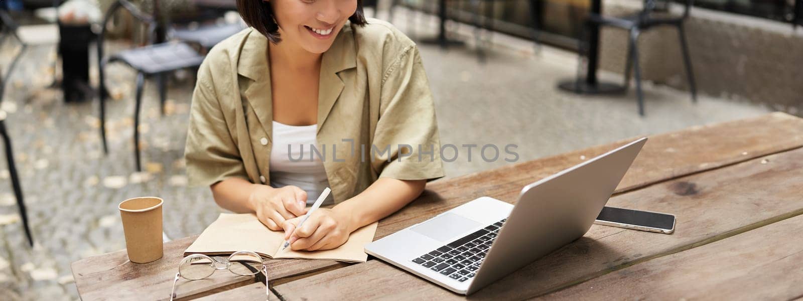 Portrait of young woman studying online, sitting with laptop, writing down, making notes and looking at computer screen, sitting in cafe outdoors.