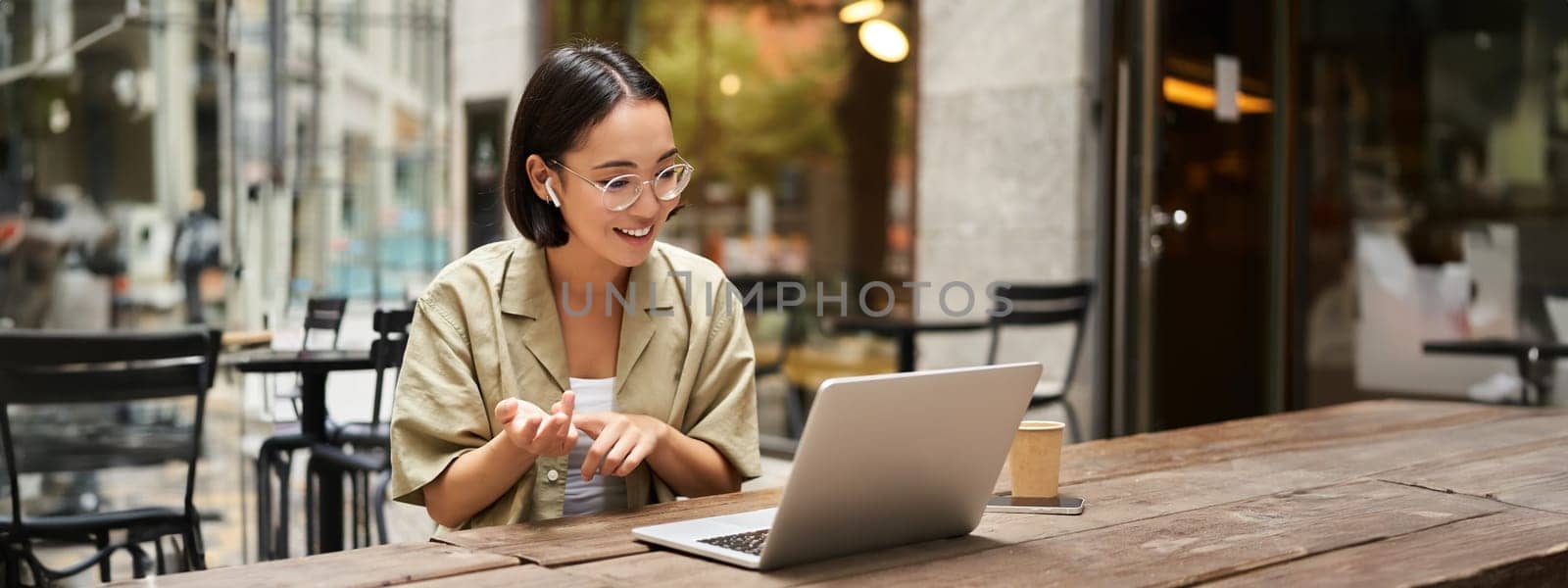 Young woman sitting on online meeting in outdoor cafe, talking to laptop camera, explaining something, drinking coffee. Digital nomad