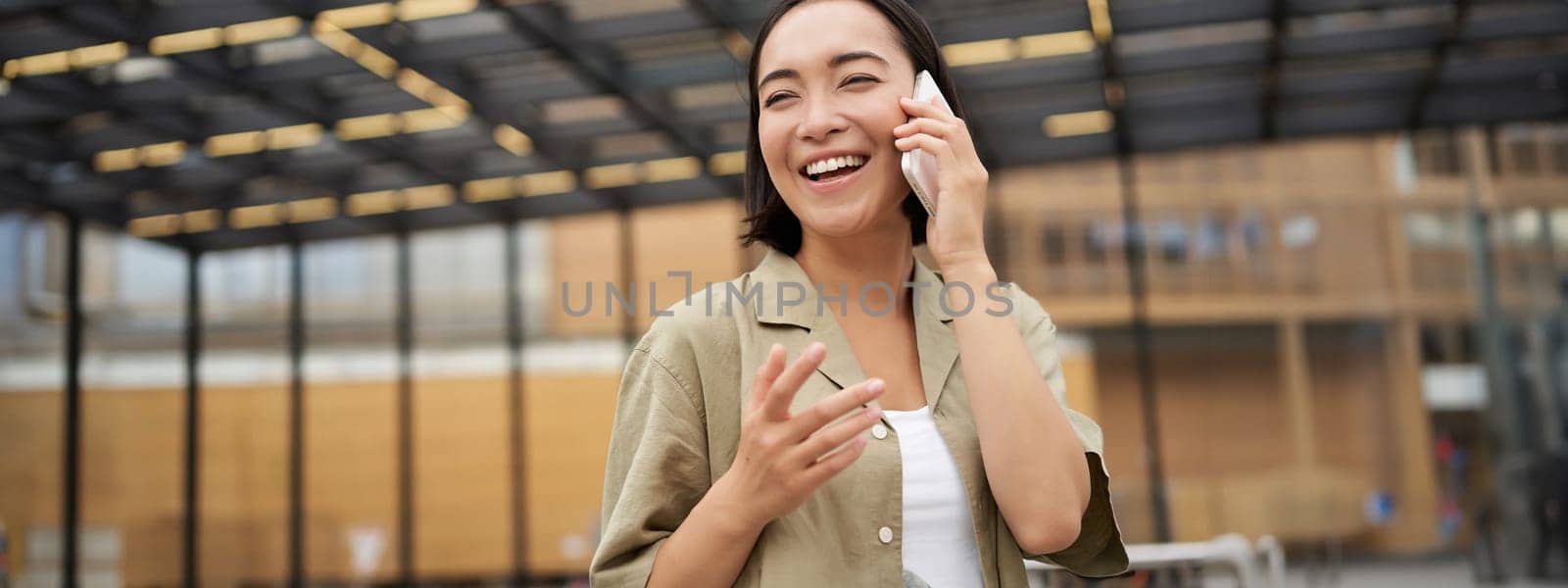 Young asian woman smiles, talks on mobile phone and walks in city centre, poses outdoors by Benzoix