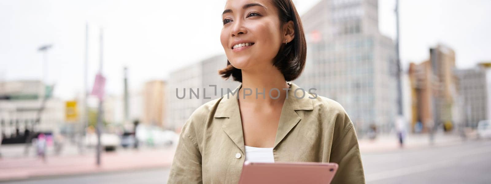 Portrait of asian girl smiles, reads on tablet, uses device while stands on city street.