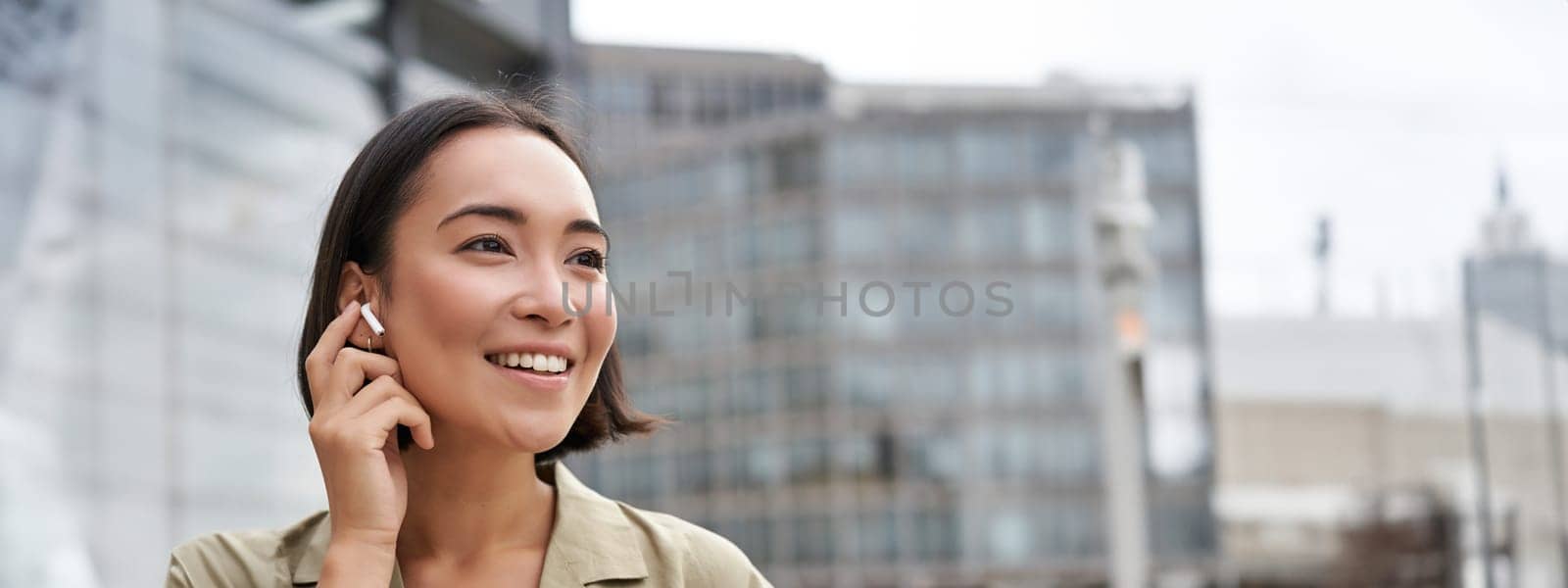 Vertical shot of smiling asian woman in wireless headphones, enjoys listening to music in earphones, holds mobile phone.