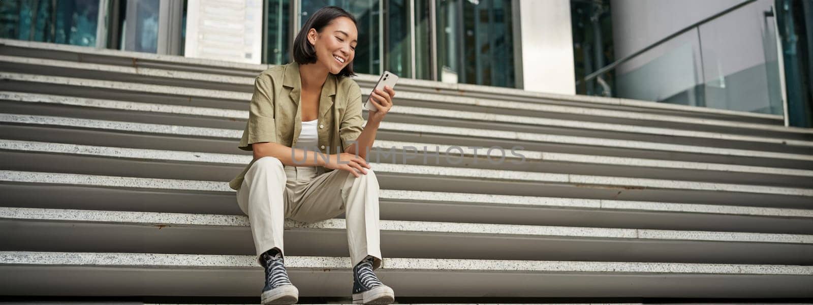 Portrait of happy smiling asian woman, sitting outdoors near building, using smartphone. Technology concept by Benzoix