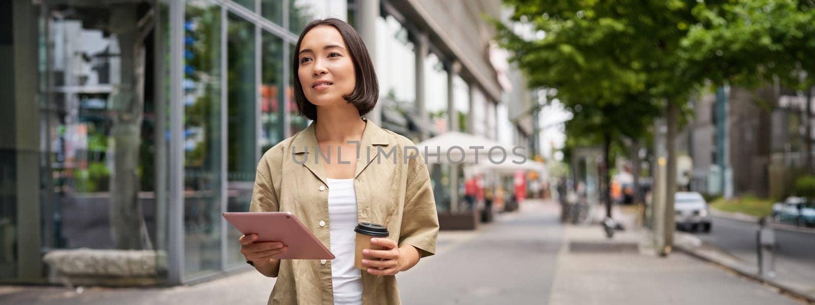 Portrait of smiling young woman walking in city with tablet, drinking takeaway coffee, going down the street with happy expression.