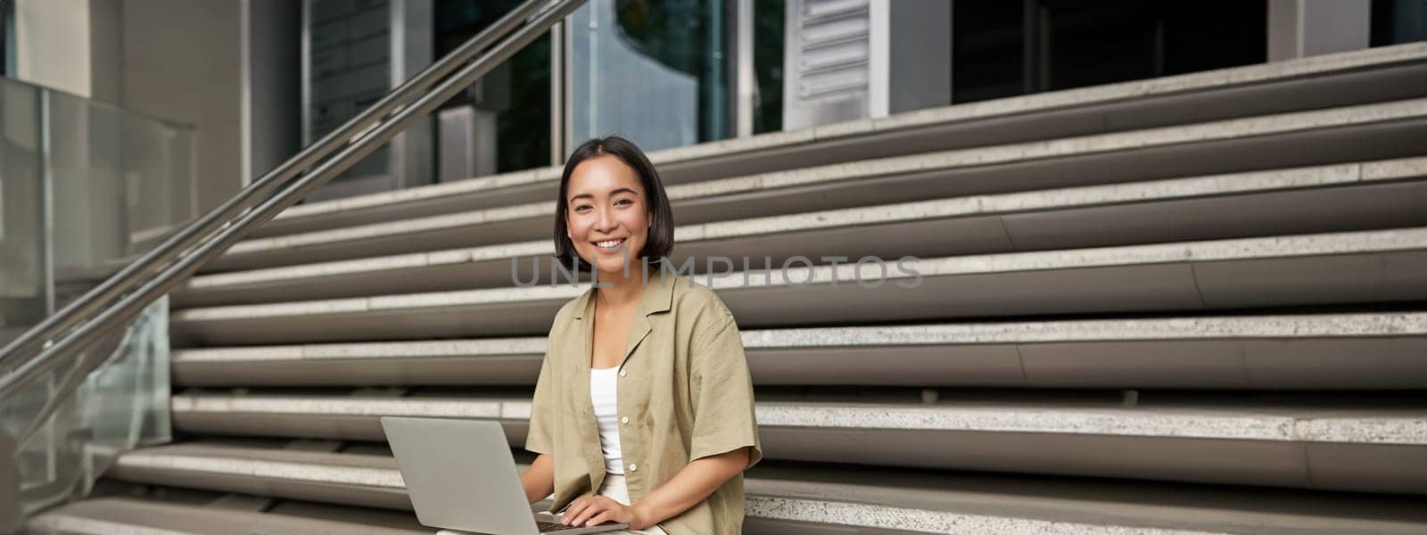 Vertical shot of asian girl sits with laptop, drinks coffee on university stairs. Young woman, student does her homework outdoors by Benzoix