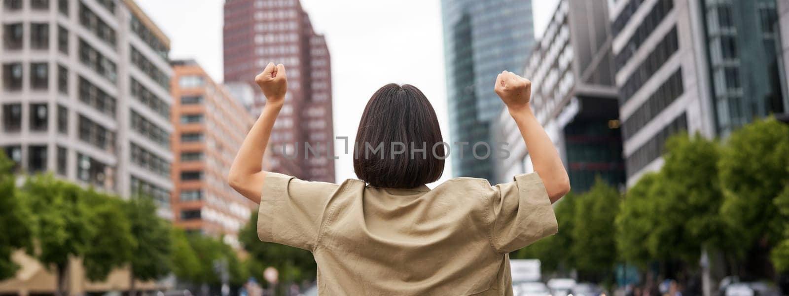 Excitement. Happy girl rear view, raising her hands up and looking at big city skyscrappers, feeling confident, cheering and triumphing by Benzoix