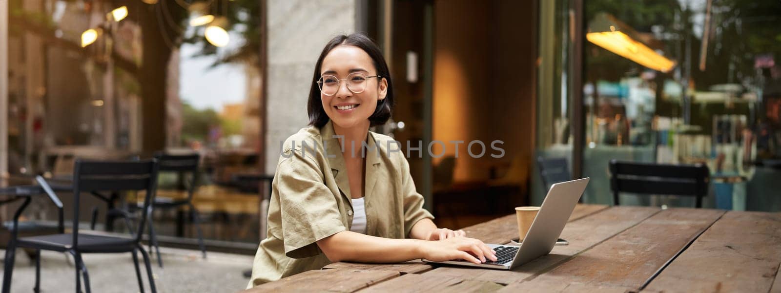 Portrait of asian girl works in cafe, uses laptop and sits outdoors on street. Digital nomad and online learning concept.