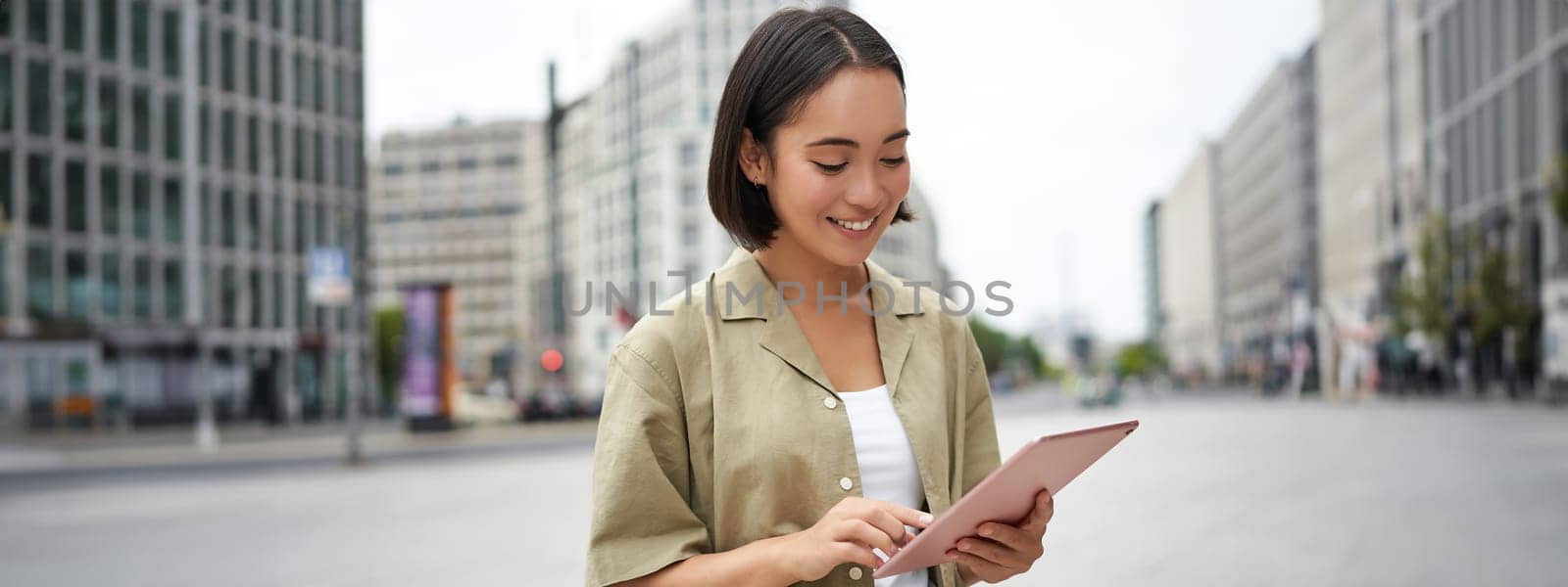 Young smiling asia woman with tablet, standing on street in daylight.